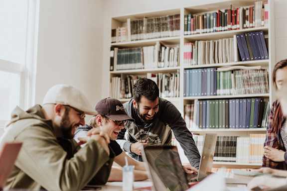A group of people are gathered around a laptop in a room with bookshelves in the background.