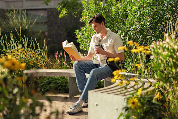 A man is sitting on a bench reading a book and holding a phone.