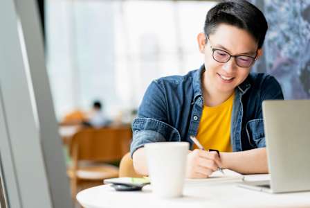 A person in a yellow shirt is writing on a piece of paper while sitting at a table with a laptop and a cup.