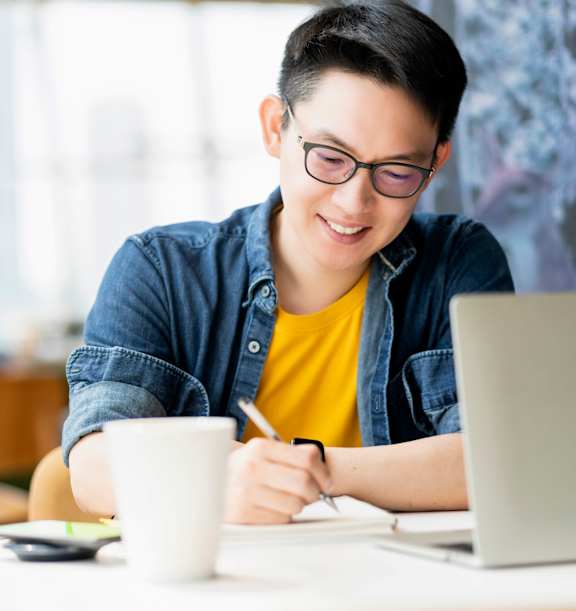 A person in a yellow shirt is writing on a piece of paper while sitting at a table with a laptop and a cup.
