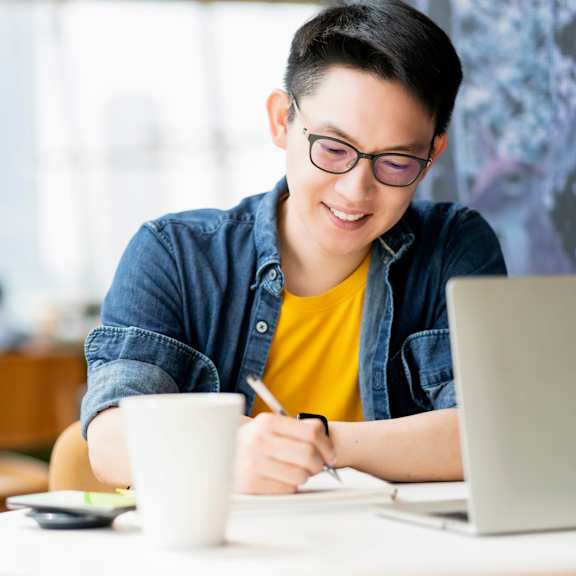 A person in a yellow shirt is writing on a piece of paper while sitting at a table with a laptop and a cup.