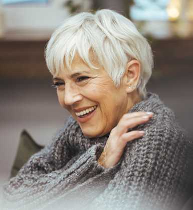 A woman with short white hair is wearing a grey sweater.