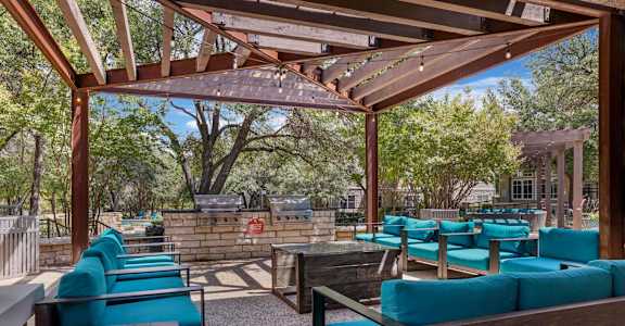 A wooden pergola with blue cushioned seating is surrounded by trees.