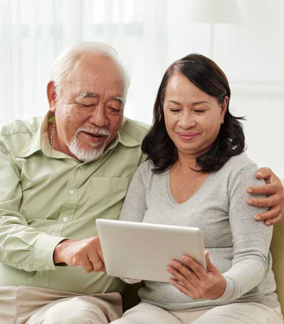 An elderly man and woman are sitting on a couch looking at a tablet.at Rosswood Villa Apartments, Santa Ana California