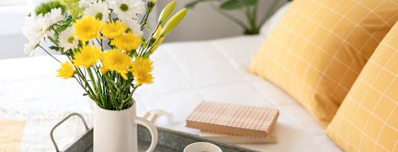 A tray with a mug, a cup of coffee, a vase with flowers and a book on a bed.