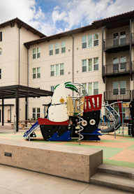 A playground area with a slide and a teddy bear slide in front of a building.