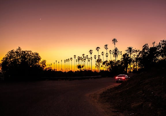 A car is driving down a road with palm trees on the side and a sunset in the background.