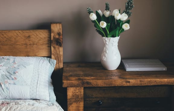 a white vase with flowers on a table next to a bed