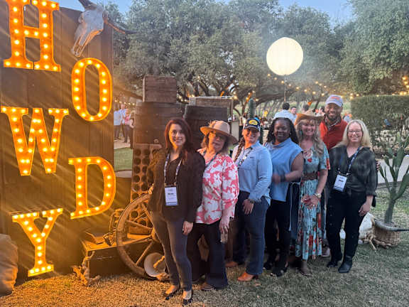A group of people are standing in front of a sign that says "HOW DY".