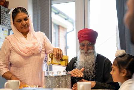 A woman in a pink shawl is serving tea to a man in a red turban and a young girl at a table.