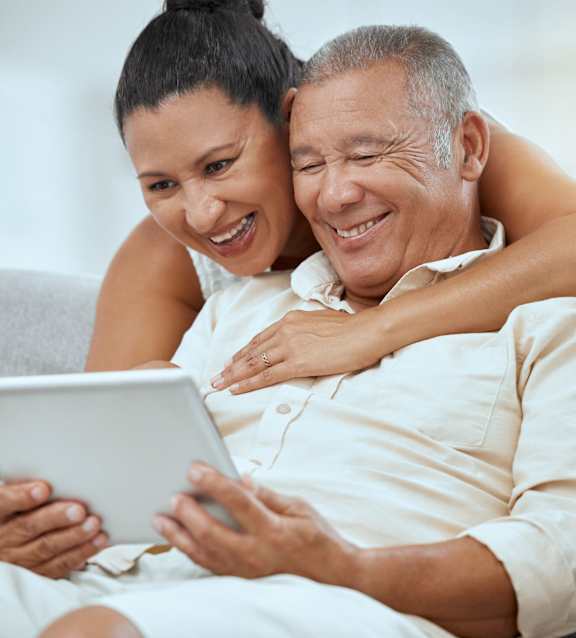 A man and woman are sitting on a couch, with the woman leaning on the man's shoulder while he holds a tablet.at The Villas 55+ II Hesperia, Hesperia, CA