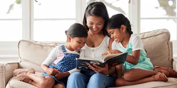 A woman and two young girls are sitting on a couch reading a book together.