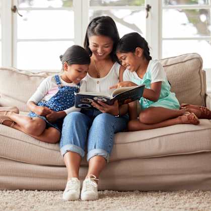 A woman and two young girls are sitting on a couch reading a book together.