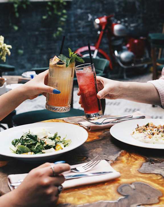 Two people toasting with drinks and plates of food on a table.