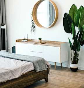 A round mirror hangs on a white wall above a wooden dresser with a bed in front of it.