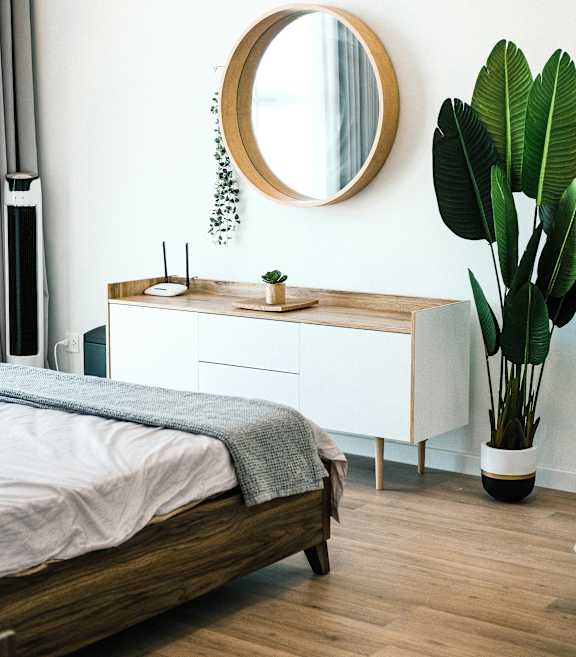 A round mirror hangs on a white wall above a wooden dresser with a bed in front of it.