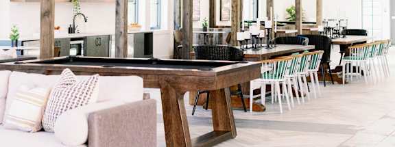 View of the market-style resident lounge with a billiards table, wooden cross beaming on the ceiling, and large pendant lighting at The Merchant in Charleston, 29403