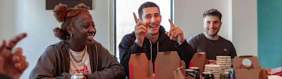 Three people sitting at a table with boxes and cans in front of them.