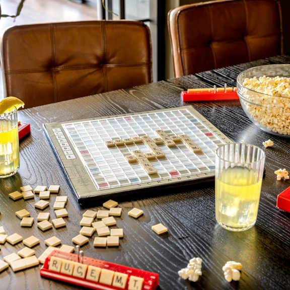 A Scrabble board game is set up on a table with a bowl of popcorn and two glasses of a yellow beverage.