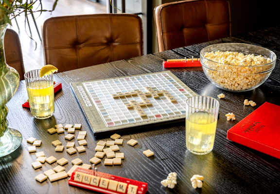 A Scrabble board game is set up on a table with a bowl of popcorn and two glasses of a yellow beverage.
