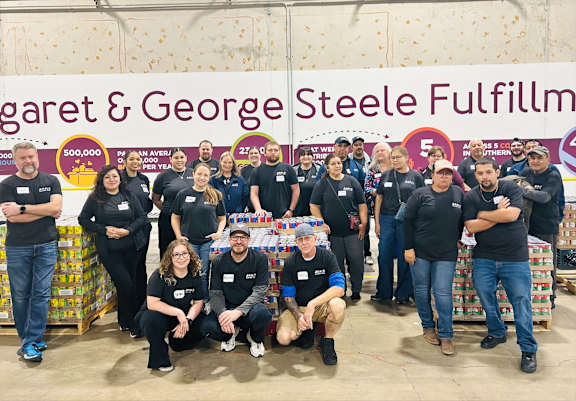 A group of people in black shirts are posing for a photo in front of a banner for Margaret & George Steele Fulfillment.