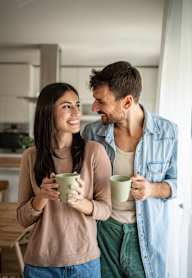 A man and a woman are standing close together, each holding a mug, in a kitchen setting.