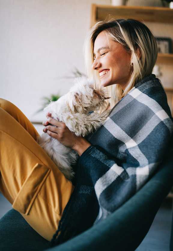 A woman is sitting on a chair holding a dog wrapped in a blanket.