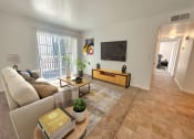 Thumbnail 4 of 17 - Bridgeside Landing Apartments in Taylorsville, Utah, Living area with tan couch, flatscreen television on wall, balcony off the room, hallway back to bedroom.
