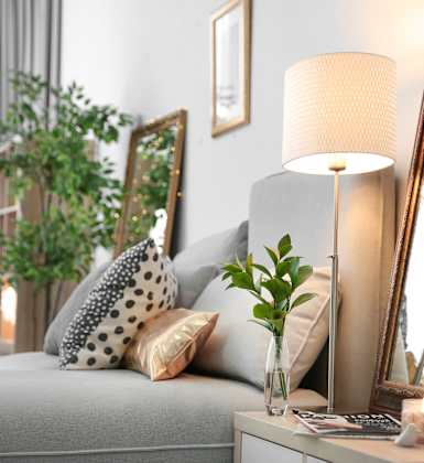 A grey sofa with a white cushion and a black and white pillow is in front of a grey wall with a framed mirror and a lamp on a table.