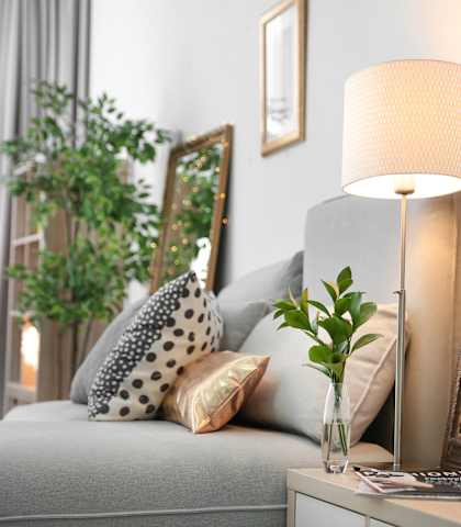 A grey sofa with a white cushion and a black and white pillow is in front of a grey wall with a framed mirror and a lamp on a table.