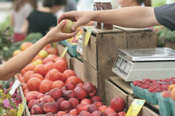 A person is handing a red apple to another person at a fruit stand.