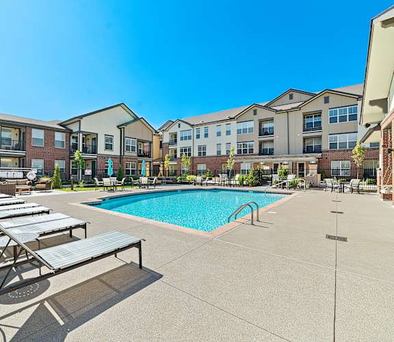 A swimming pool surrounded by sun loungers in front of apartment buildings.