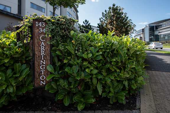 a sign for the entrance of a building with a bush with green plants