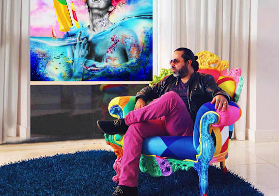 A man in a black leather jacket and pink pants is sitting on a colorful chair in front of a painting.