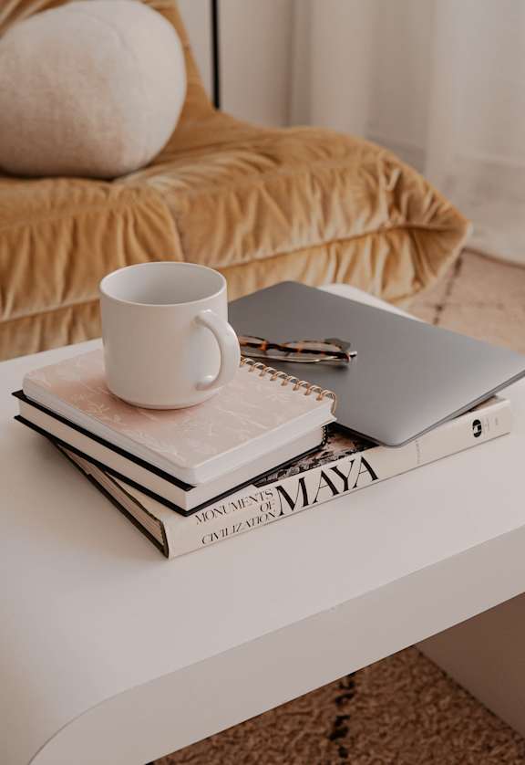 White coffee table with books, journals, a mug, a computer, and glasses on top of it. Behind the coffee table is a tan leather couch.