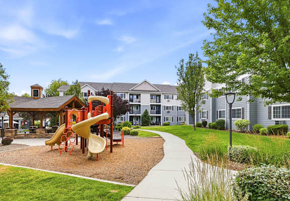 A playground with a slide and a wooden structure is surrounded by a grassy area and a sidewalk.