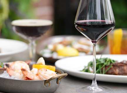 A glass of red wine is in the foreground with food in the background.