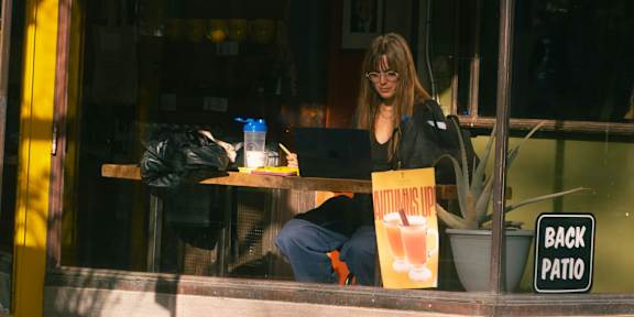 A woman is sitting at a table outside a cafe with a laptop and a bag.