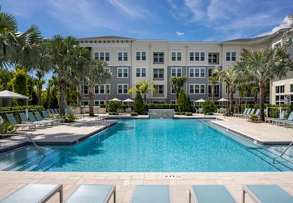 A large swimming pool surrounded by palm trees and lounge chairs in front of a multi-story building.