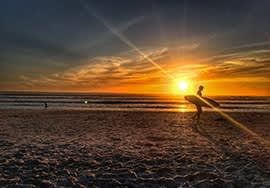 a man carrying a surfboard on the beach at sunset