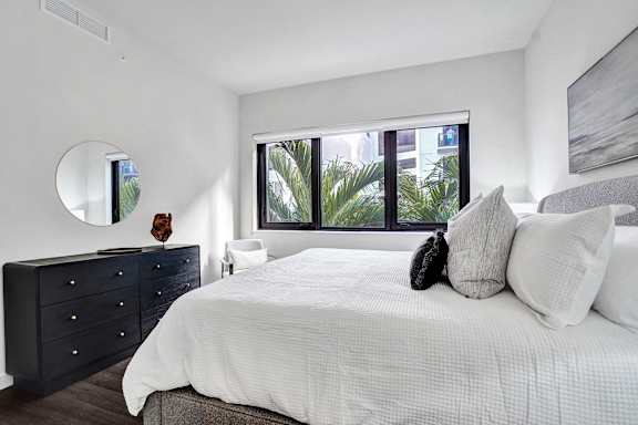 A bedroom with a large bed, a dresser, and a window overlooking a green area.