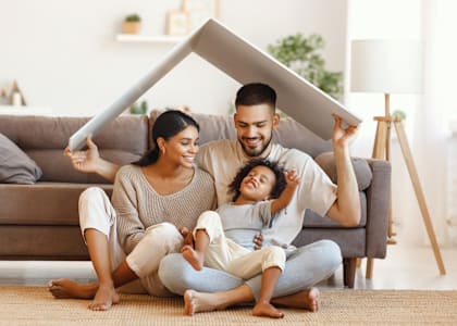 A family of three is sitting on the floor in a living room, holding a large white object.