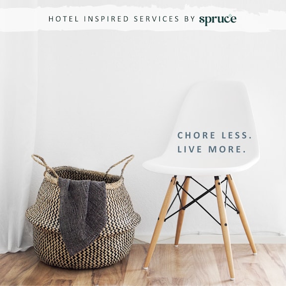 A wicker laundry basket with a grey towel in it and a white chair with wooden legs and a sign on it that says chore less live more.