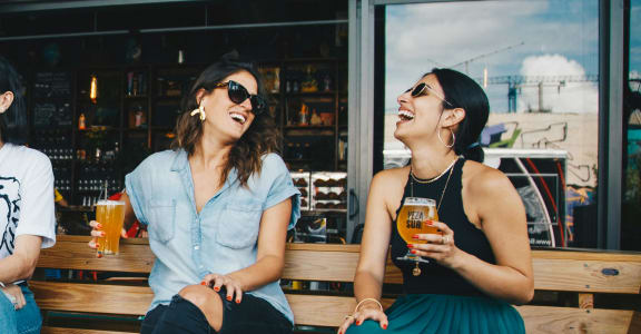 two women sitting on a bench laughing and drinking beer