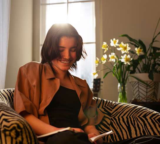 A woman is sitting on a striped couch holding a book and smiling.