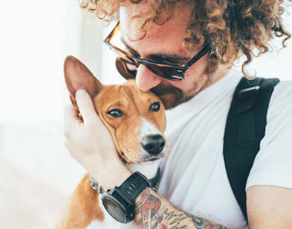 A man with curly hair and tattoos is holding a small dog close to his face.