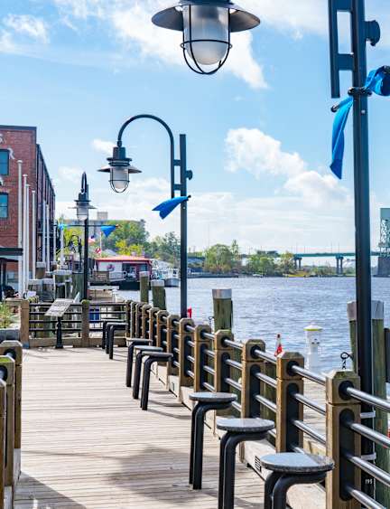 A wooden pier with benches and a lamp post overlooking a river with a bridge in the distance.