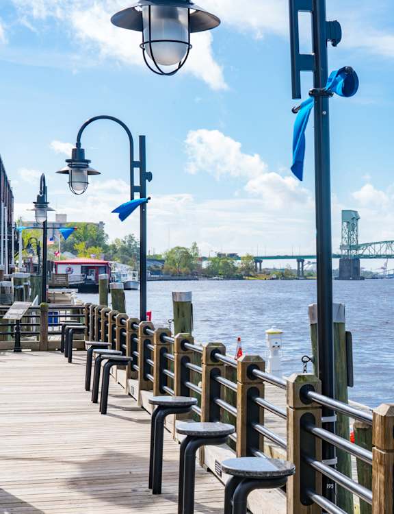 A wooden pier with benches and a lamp post overlooking a river with a bridge in the distance.