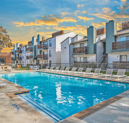 A swimming pool surrounded by lounge chairs and buildings in the background.