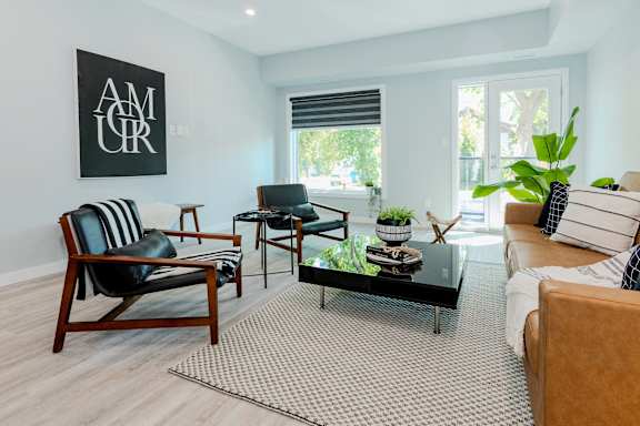 A living room with a black and white striped chair, a wooden chair, a glass coffee table, and a couch with a throw pillow.